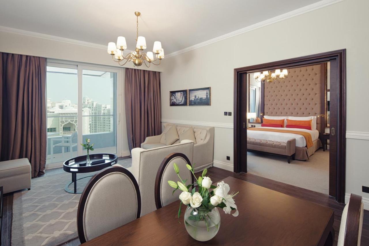 Dukes The Palm, A Royal Hideaway Hotel Dubaj Zewnętrze zdjęcie The photo depicts a well-furnished hotel suite or living space. In the foreground, there is a dining table with plush chairs and a vase of flowers. Beyond that, you can see a cozy living area with a sofa set and a coffee table, illuminated by a chand