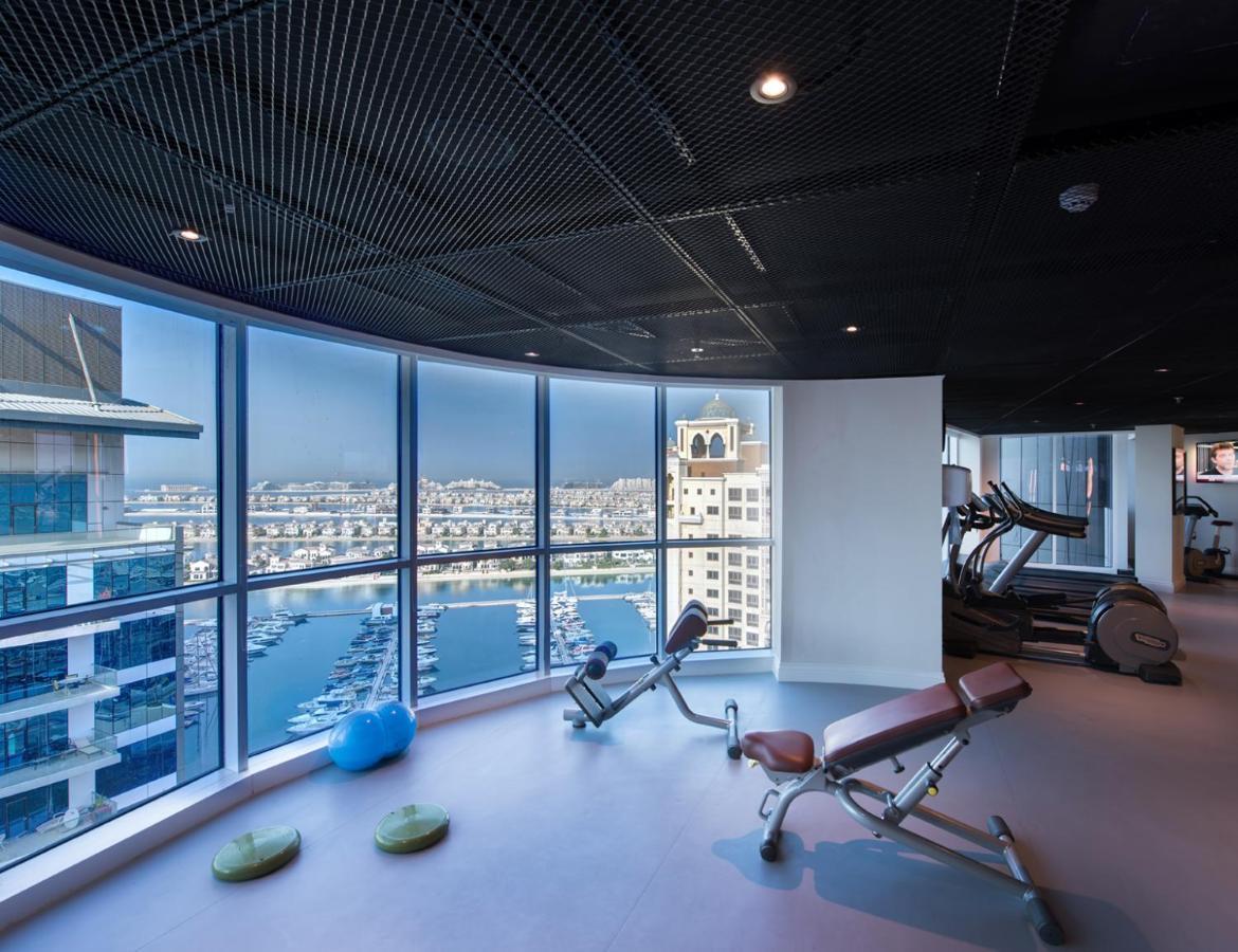 Dukes The Palm, A Royal Hideaway Hotel Dubaj Zewnętrze zdjęcie The photo shows a modern gym with large windows offering a panoramic view of a marina or waterfront area. Inside, there are exercise machines, including a couple of benches for weight training. The flooring is smooth, and there are a few exercise acc