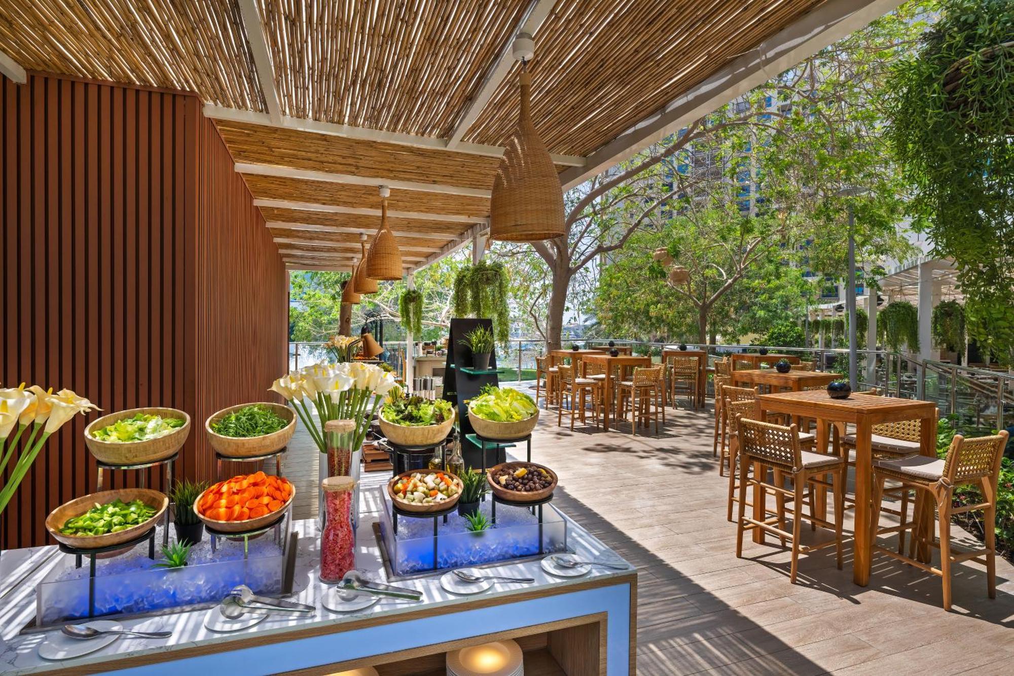 Dukes The Palm, A Royal Hideaway Hotel Dubaj Zewnętrze zdjęcie The photo showcases a bright and inviting outdoor dining space. In the foreground, there is a buffet table with various salads and fresh vegetables displayed in bowls, including items like lettuce, carrots, and other colorful ingredients, set against