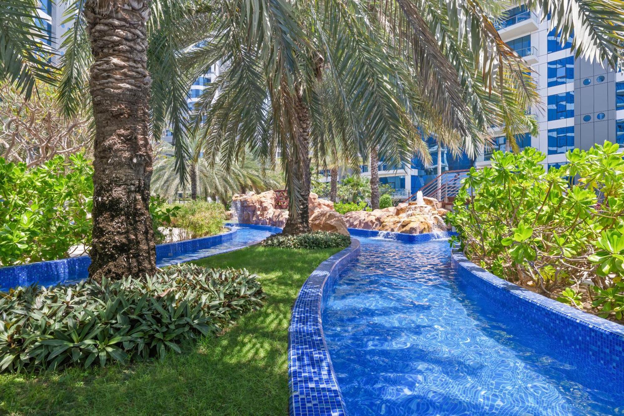 Dukes The Palm, A Royal Hideaway Hotel Dubaj Zewnętrze zdjęcie The photo depicts a beautifully landscaped tropical area featuring a winding waterway with clear blue water, bordered by lush greenery and palm trees. The vibrant blue tiles lining the water add a decorative touch, while the overall setting suggests 