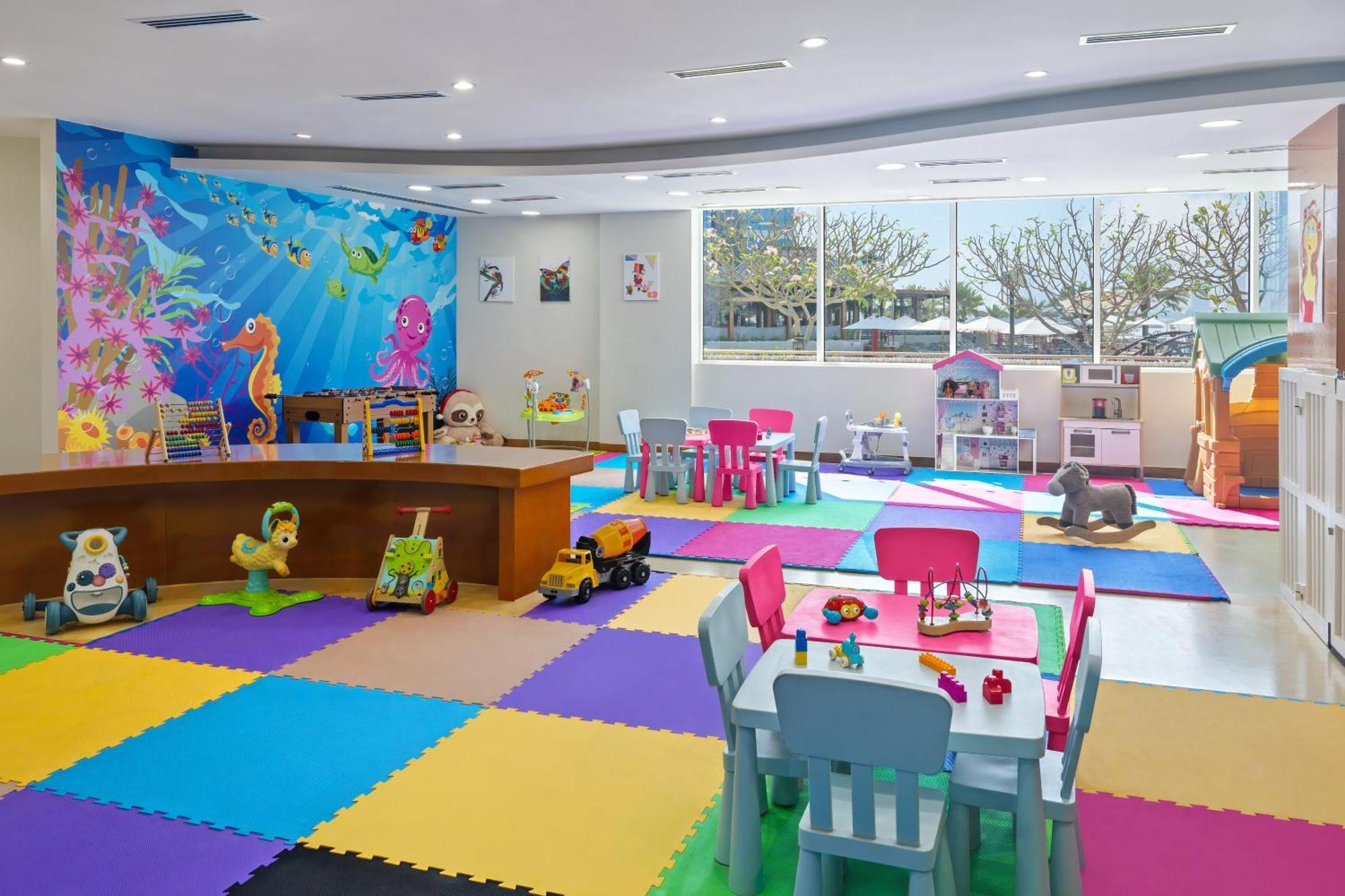 Dukes The Palm, A Royal Hideaway Hotel Dubaj Zewnętrze zdjęcie The photo shows a bright and colorful children's playroom. The floor is covered with interlocking foam mats in various colors, including blue, green, yellow, and purple. There is a large table made of wood at the center, surrounded by colorful chairs