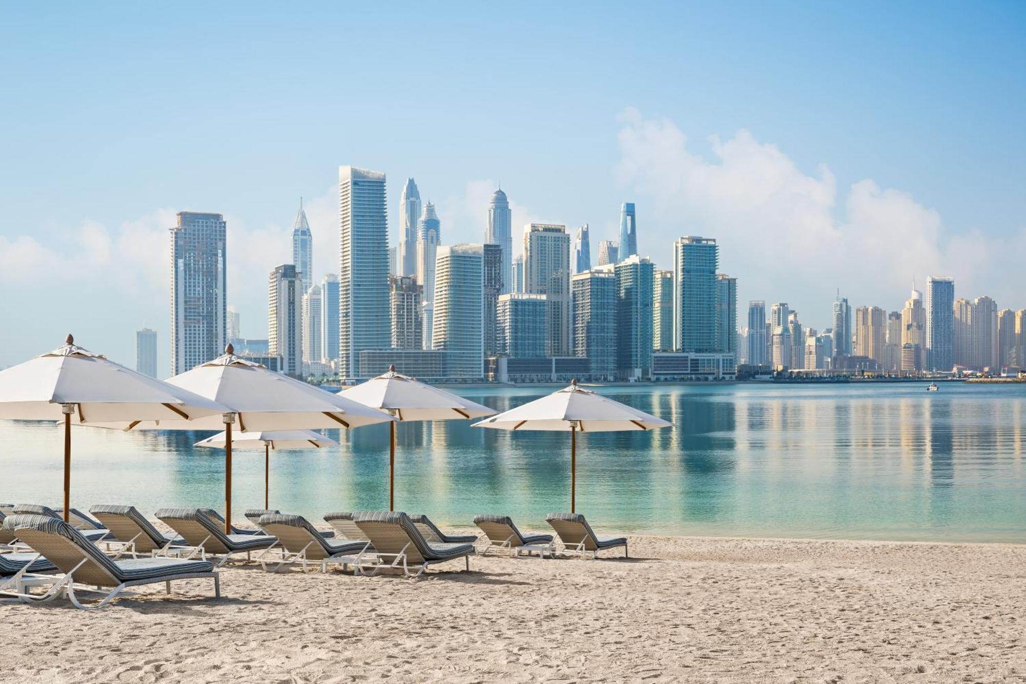 Dukes The Palm, A Royal Hideaway Hotel Dubaj Zewnętrze zdjęcie The photo showcases a serene beach scene with several lounge chairs positioned on the sandy shore. Each chair is paired with a large umbrella for shade. In the background, a modern city skyline is visible, featuring tall skyscrapers and a clear blue 