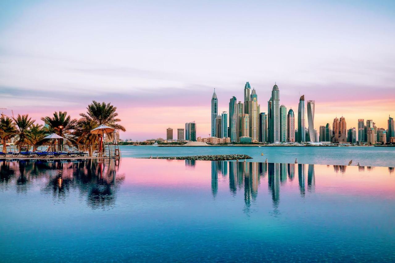 Dukes The Palm, A Royal Hideaway Hotel Dubaj Zewnętrze zdjęcie The image showcases a stunning skyline view of a modern city, likely during sunrise or sunset, as indicated by the beautiful pastel colors in the sky. The foreground features a calm body of water, reflecting the skyscrapers and the colorful sky. Lush